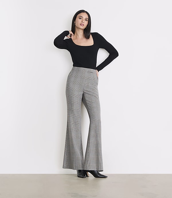 A woman in a black top and plaid bell-bottom pants stands against a plain white background.