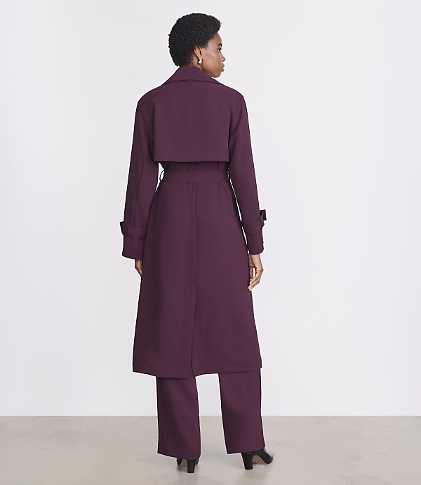 A woman in a plum trench coat and matching pants stands against a plain white background.
