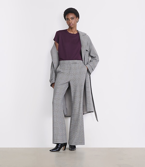 A woman in a plum t-shirt, plaid wide-leg pants, and a grey trench coat stands against a plain white background.