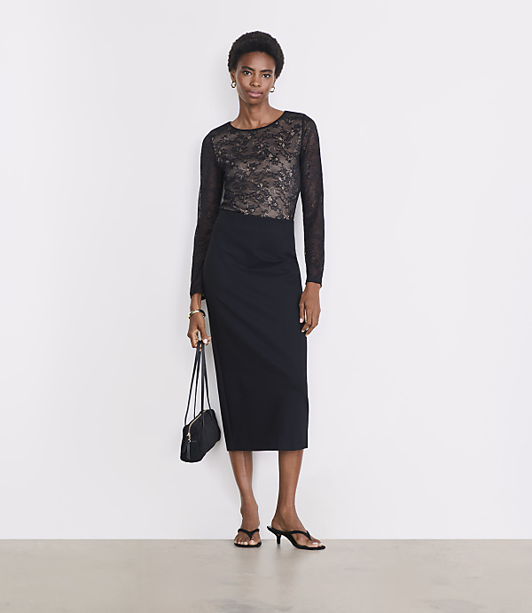 A woman in a black lace top and pencil skirt stands against a plain white background, holding a small black purse.