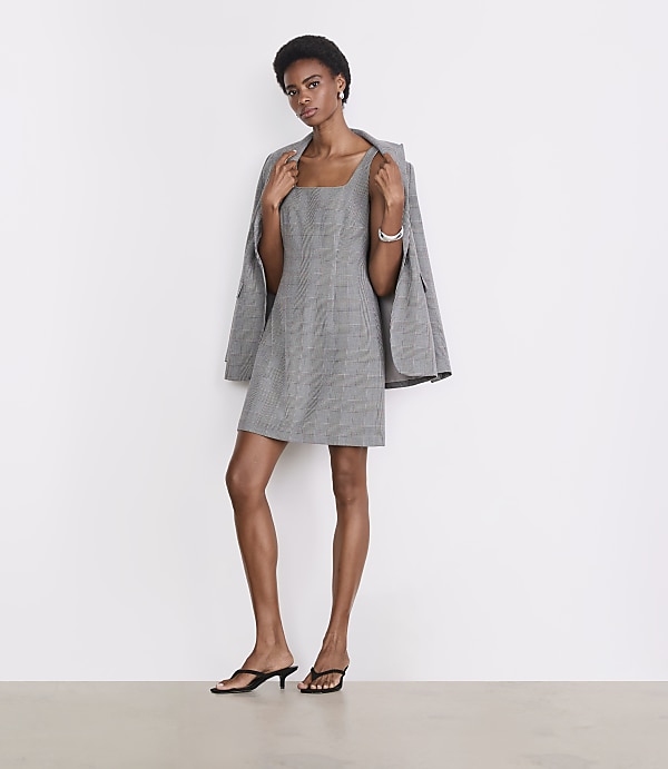 A woman in a plaid dress and heels stands against a plain white background with a blazer draped over her shoulders.