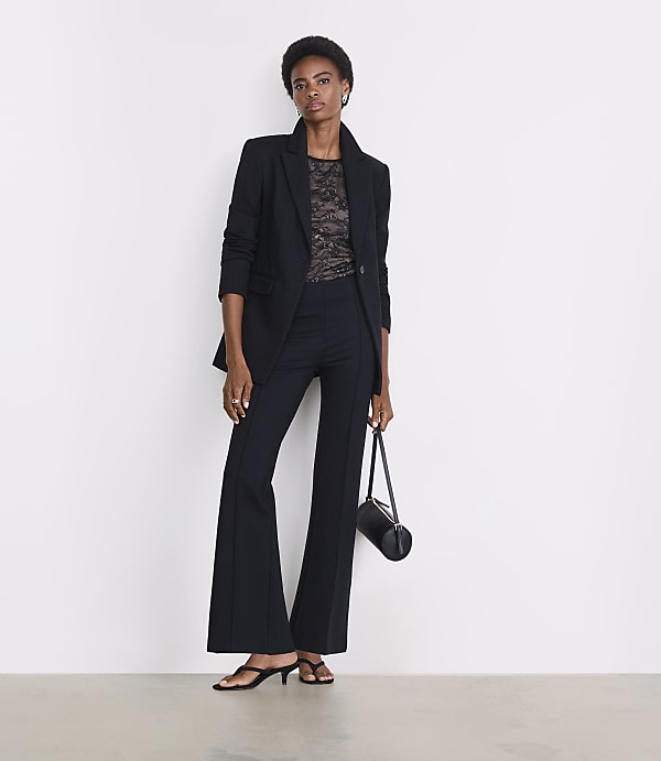A woman in a black blazer and flared pants stands against a plain white background, holding a small black bag.