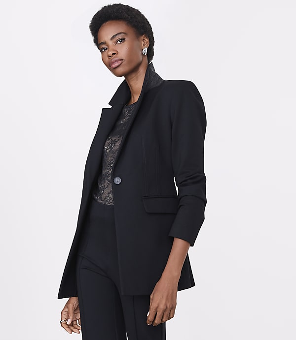A woman in a black blazer and lace top stands against a plain white background.