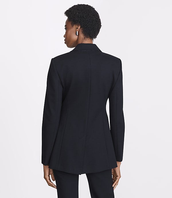 A woman in a black blazer stands against a plain white background.