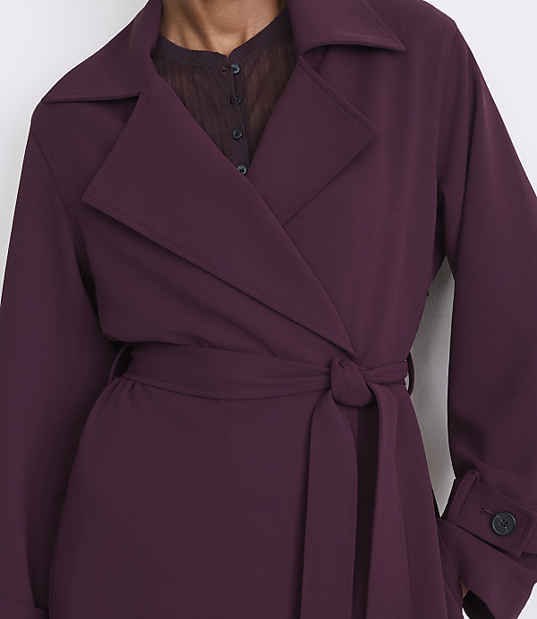 A person in a deep plum trench coat with a tie belt stands against a plain white background.