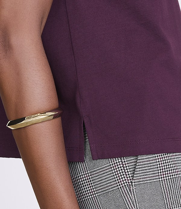 A person's arm wearing a gold bracelet is visible in the foreground, with a plum-colored shirt and plaid pants in the background.