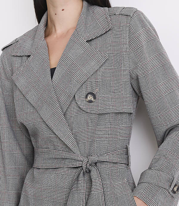 A woman in a grey plaid trench coat with a tied belt stands against a white background.