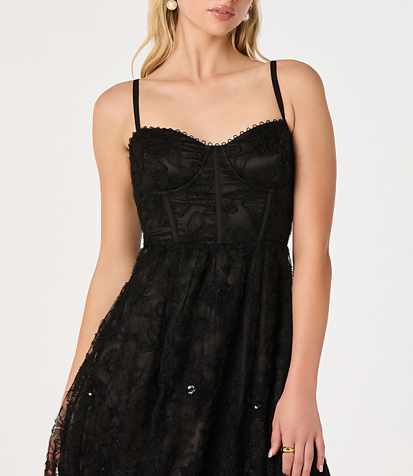 A woman in a black lace dress with a corset bodice stands against a plain white background.
