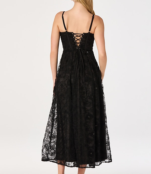 A woman in a black lace corset dress stands against a plain white background.