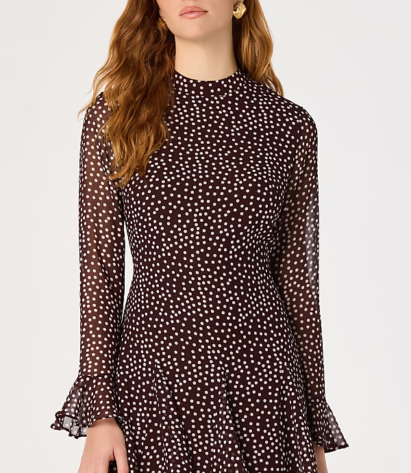 A woman in a dark, polka-dotted dress with bell sleeves stands against a plain white background.