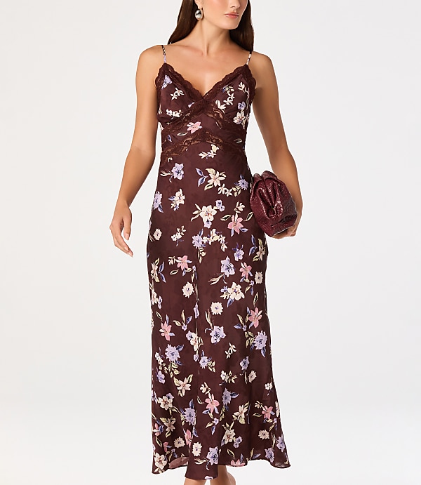 A woman in a floral slip dress and holding a clutch walks against a plain white background.