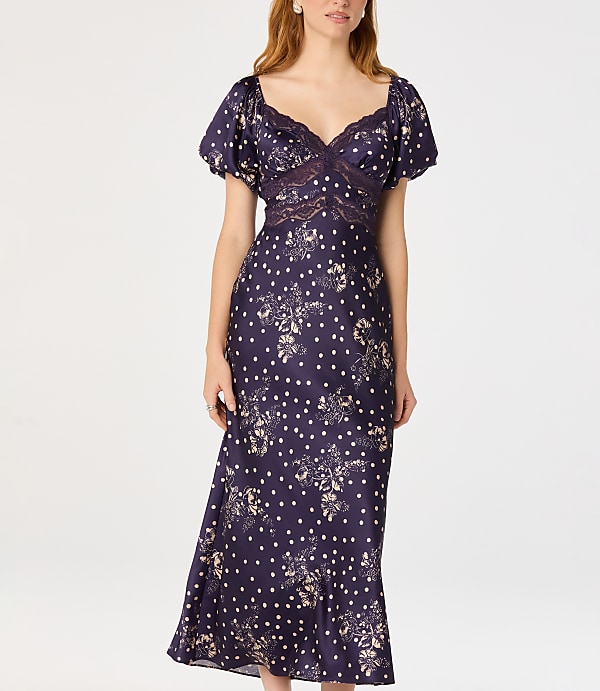 A woman in a dark floral and polka dot dress stands against a plain white background.