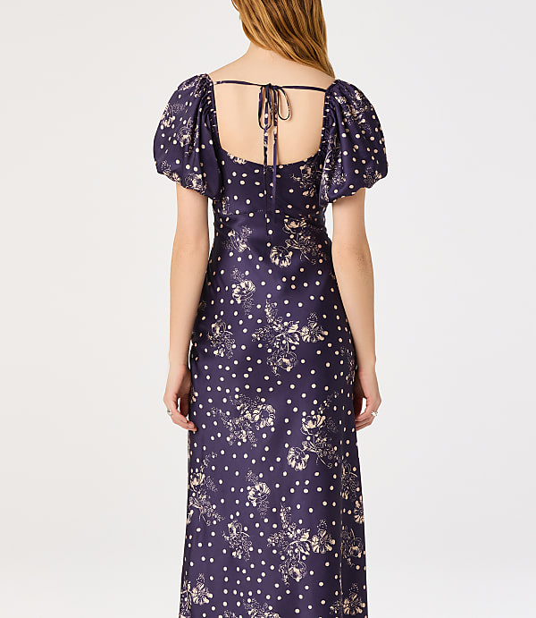A woman in a dark blue floral and polka dot dress stands against a plain white background.