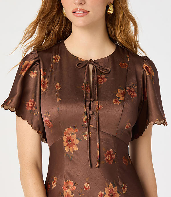 A woman with auburn hair wears a brown floral dress with flutter sleeves and a tie neck, against a plain white background.