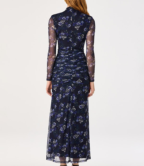 A woman in a long, dark blue floral mesh dress stands against a plain white background.