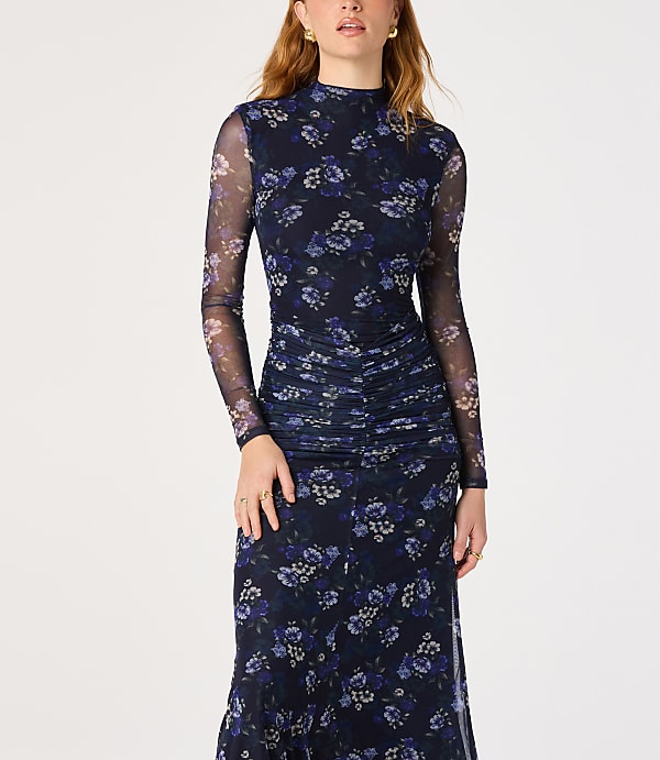 A woman in a dark floral mesh dress with ruched detailing stands against a plain white background.