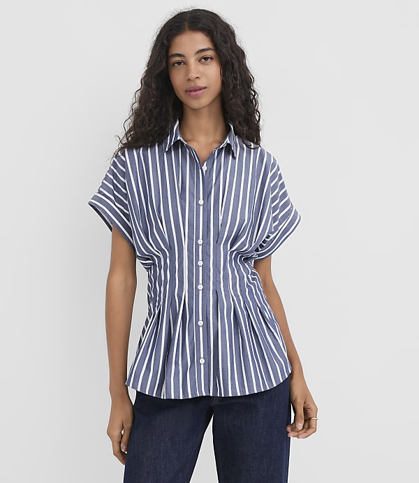A woman with dark, curly hair stands in the foreground wearing a blue and white striped shirt and dark jeans, against a plain white background.