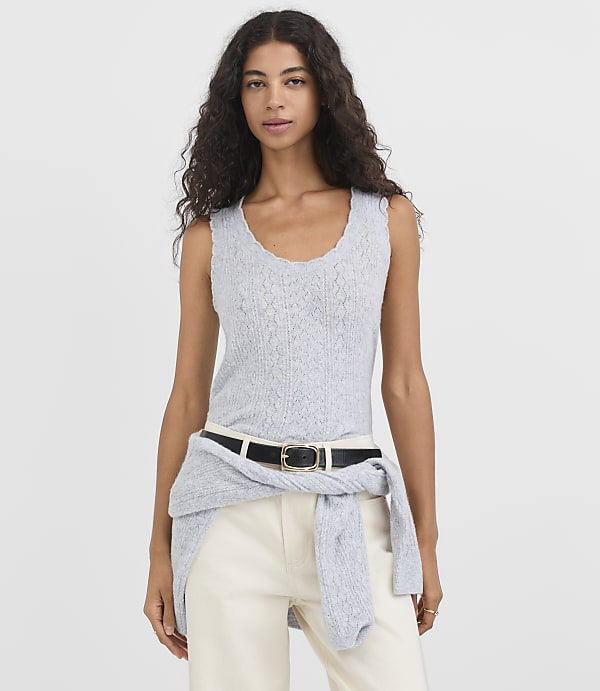 A woman in a light blue sleeveless top and cream pants stands against a plain white background, with a gray sweater tied around her waist.