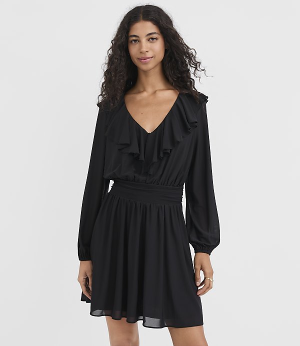 A woman in a black ruffled dress stands against a plain white background.