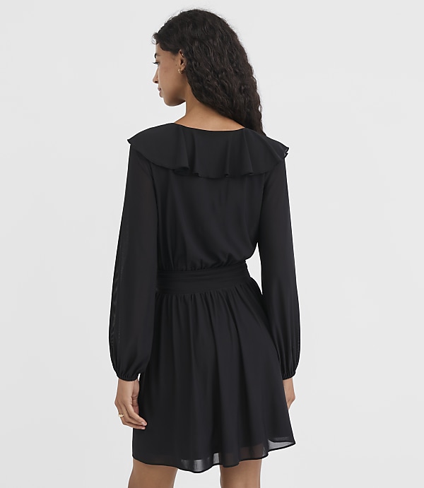 A woman in a black ruffled dress stands against a plain white background.