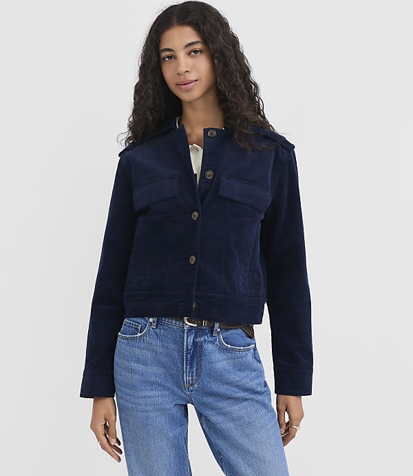A woman in a navy corduroy jacket and blue jeans stands against a plain white background.