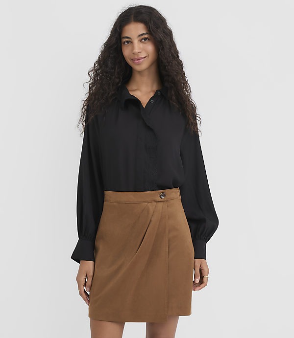 A woman with long, dark, curly hair stands in the foreground wearing a black blouse and a brown skirt, against a plain white background.