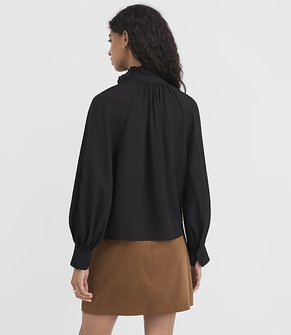A woman in a black ruffled-neck blouse and brown skirt stands against a plain white background.