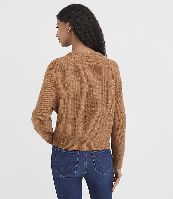 A woman in a speckled brown sweater and blue jeans stands against a plain white background.