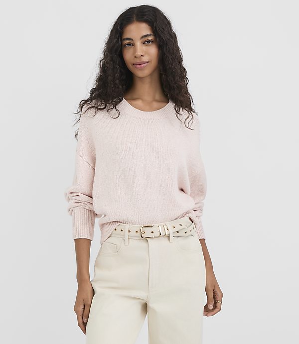 A woman in a light pink sweater and cream pants stands against a plain white background.