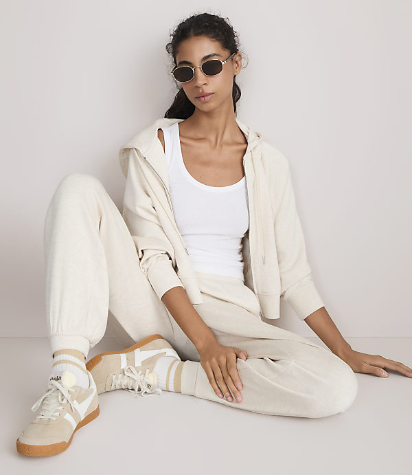 A woman in a white tank top, cream hoodie, and matching sweatpants sits on a light-colored floor, her legs crossed and wearing sneakers, against a plain, light-colored background.