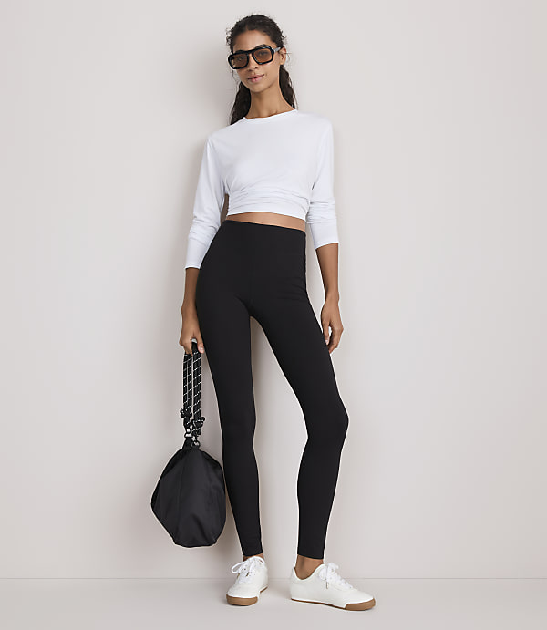 A woman in a white long-sleeved crop top and black leggings stands against a plain white background, holding a black bag.