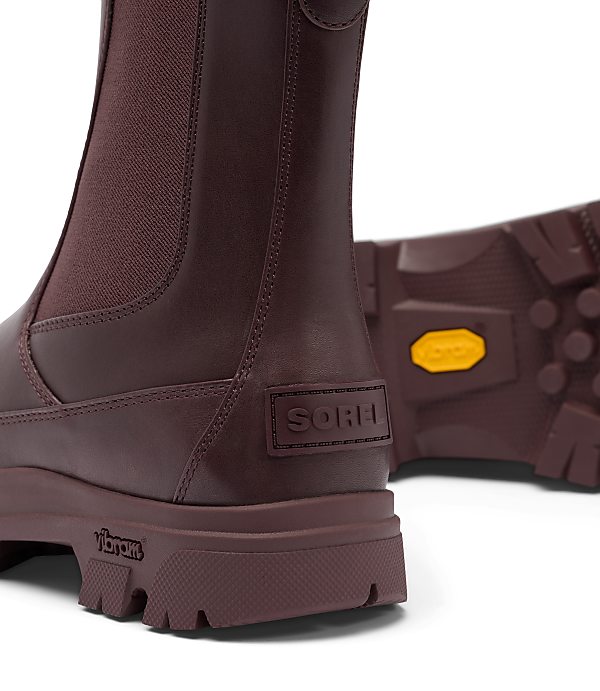 A close-up of burgundy leather Chelsea boots with thick soles and a Vibram logo is set against a white background.