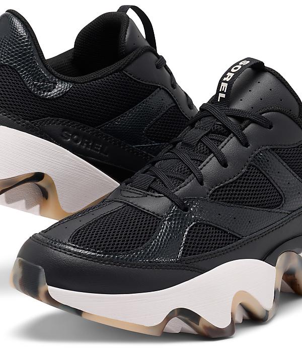A pair of black Sorel sneakers with white soles and a marbled pattern on the heel and sole are in the foreground, with a blurred background.