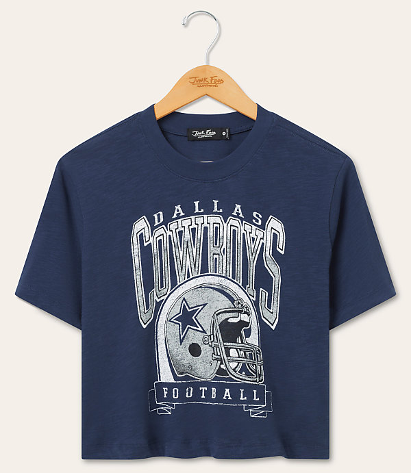 A navy blue Dallas Cowboys t-shirt with a graphic of a helmet and team name hangs on a wooden hanger against a plain beige background.