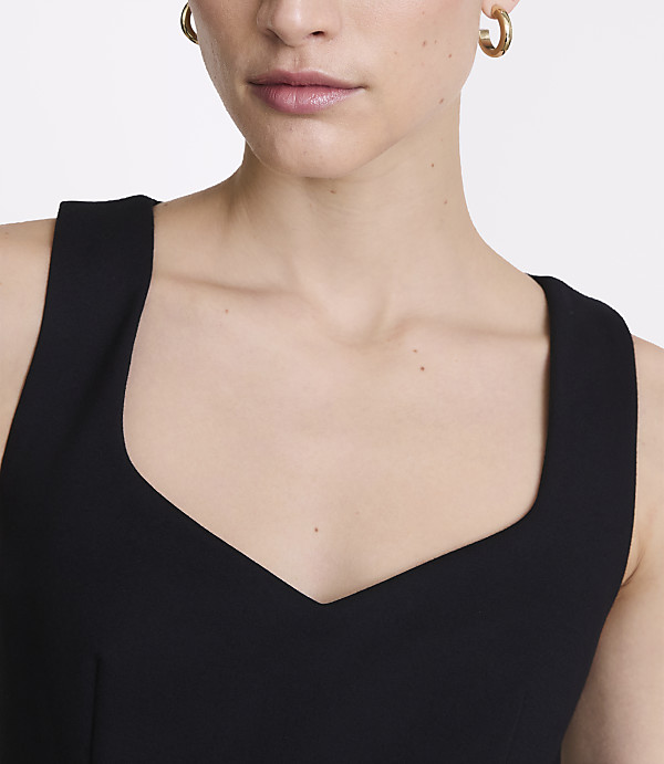 A woman with gold hoop earrings wears a black dress with a sweetheart neckline, against a plain white background.
