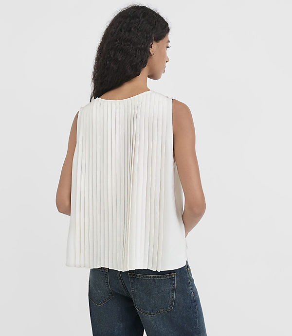A woman in a white pleated top and blue jeans stands against a plain white background.