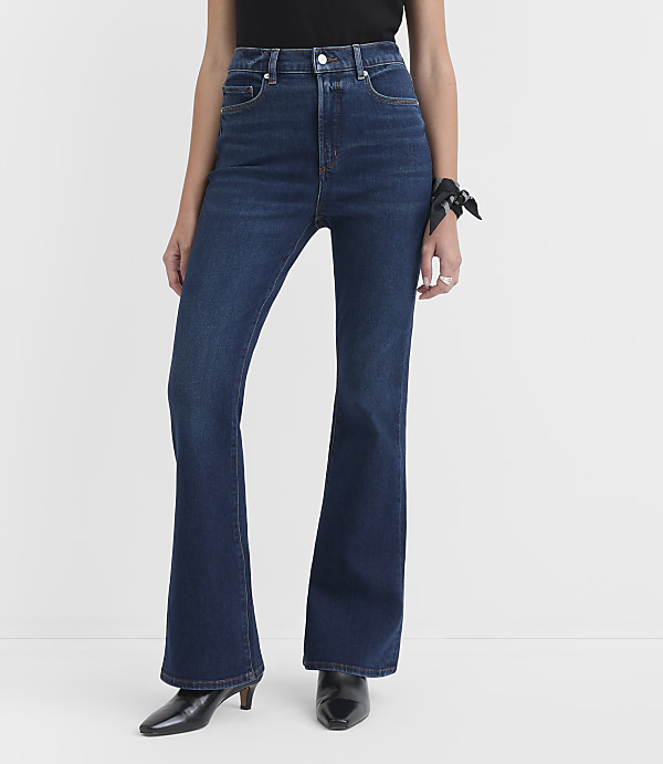 A person wearing dark wash flare jeans and black heels stands against a plain white background.