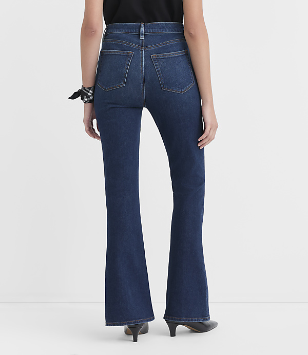 A person wearing dark wash flare jeans and black heels stands against a plain white background.