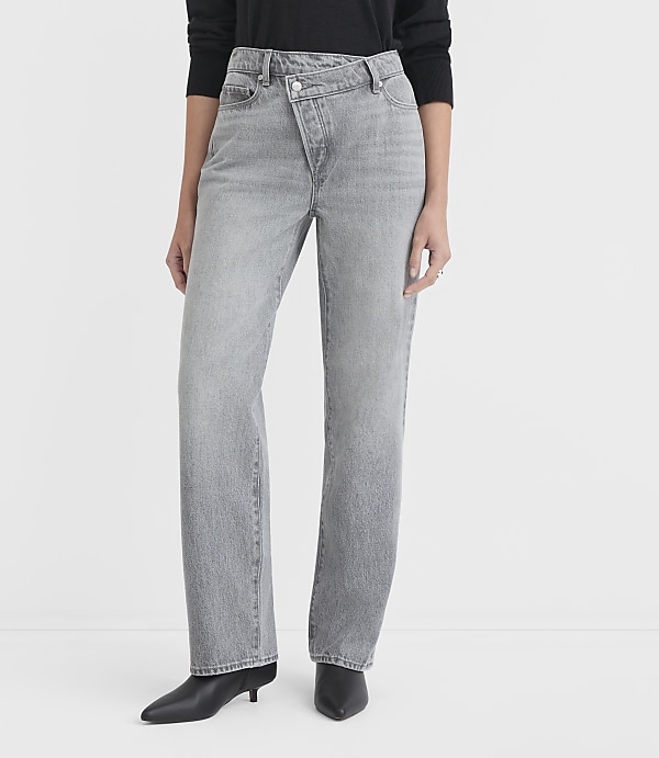 A person stands in light grey jeans with a unique criss-cross waistband and black pointed-toe heels against a plain white background.