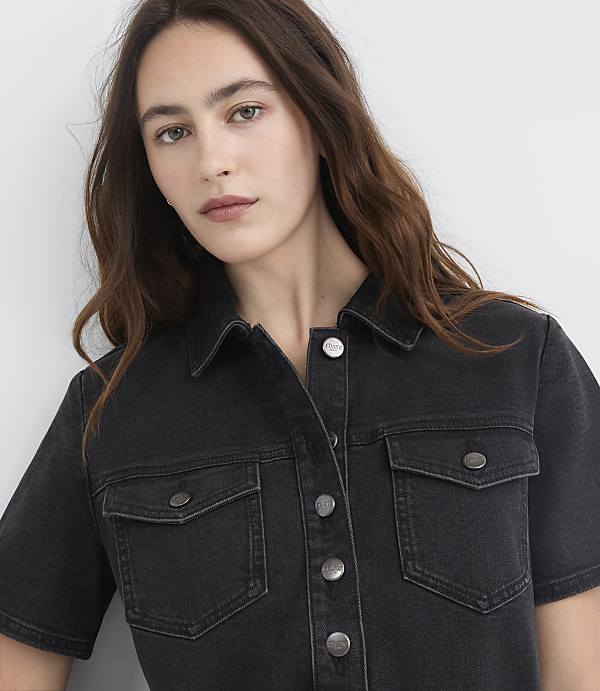 A woman with long brown hair wears a dark denim shirt with two chest pockets, set against a plain gray background.