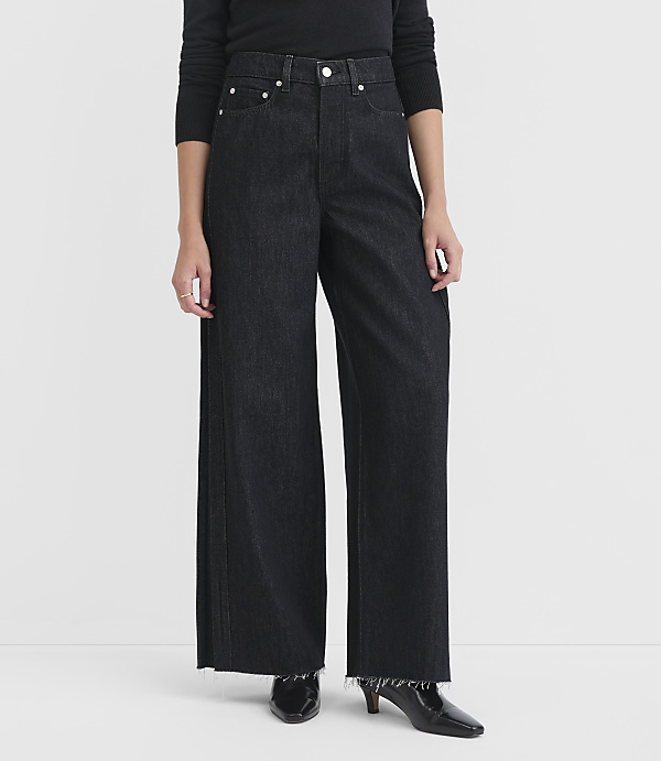 A person stands in dark wash wide-leg jeans with frayed hems and black heeled boots against a plain white background.