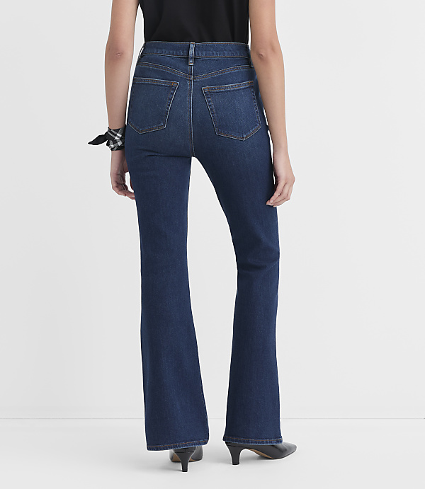 A person wearing dark wash flare jeans and black heels stands against a plain white background.