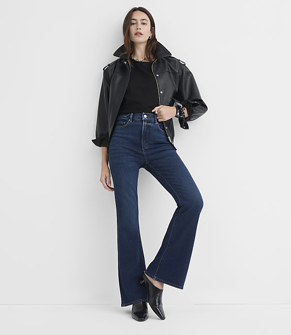 A woman in a black leather jacket and dark wash flare jeans stands against a plain white background.