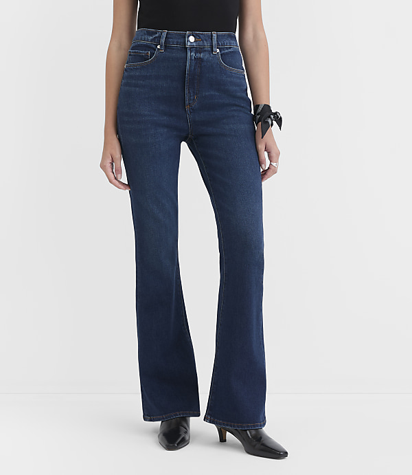 A person stands in dark wash flare jeans and black heels against a plain white background.