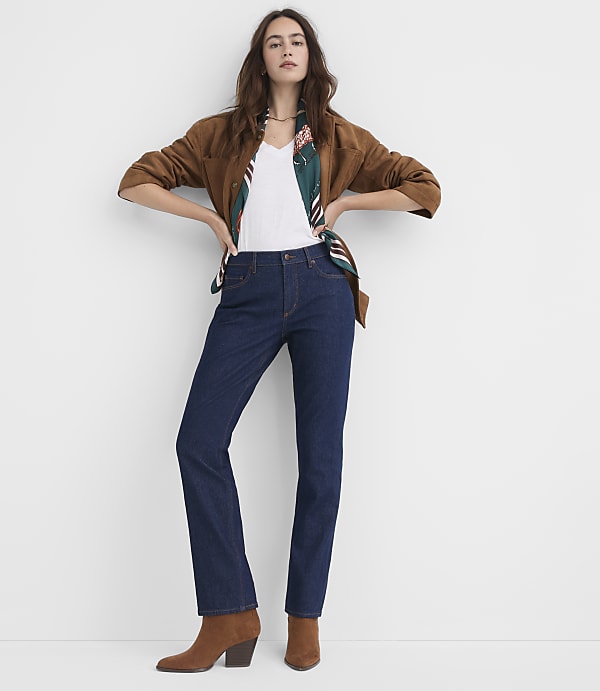 A woman in a brown jacket, white top, and dark jeans stands against a plain white background.