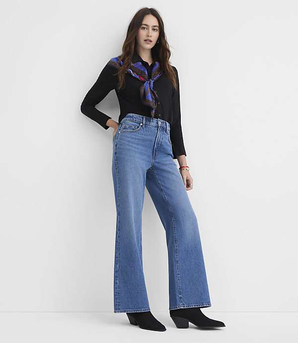 A woman in a black shirt and wide-leg jeans stands against a plain white background.