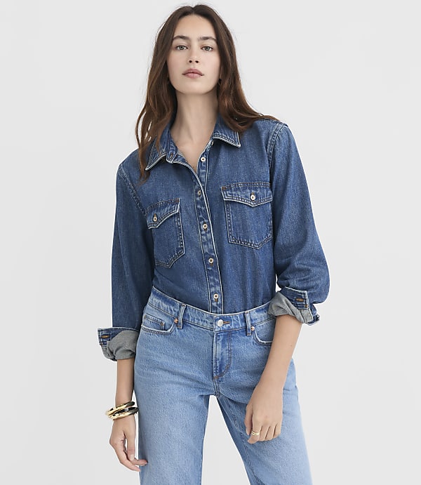 A woman in a denim shirt and jeans stands against a plain white background.