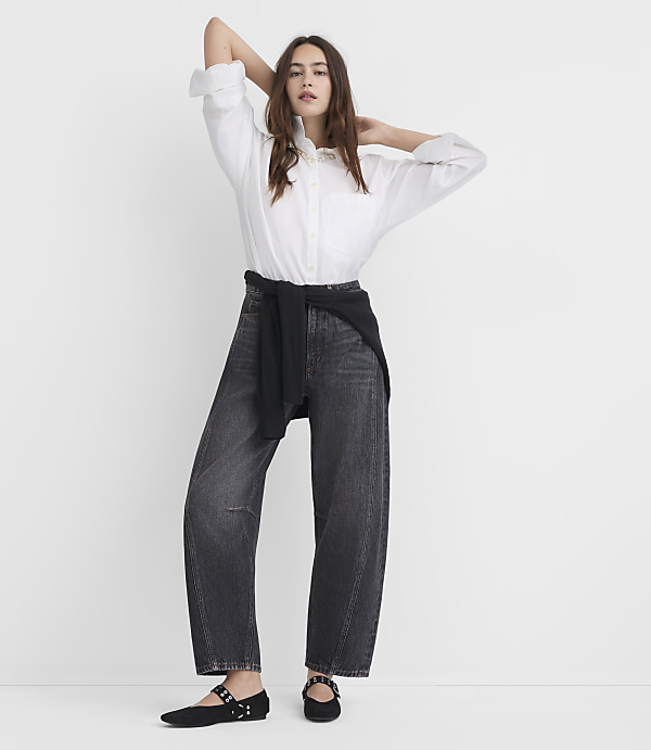 A woman in a white blouse and dark jeans stands with her arms behind her head against a plain white background.