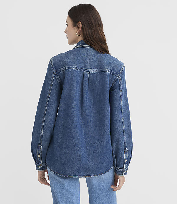 A woman in a denim shirt and jeans stands against a plain white background.