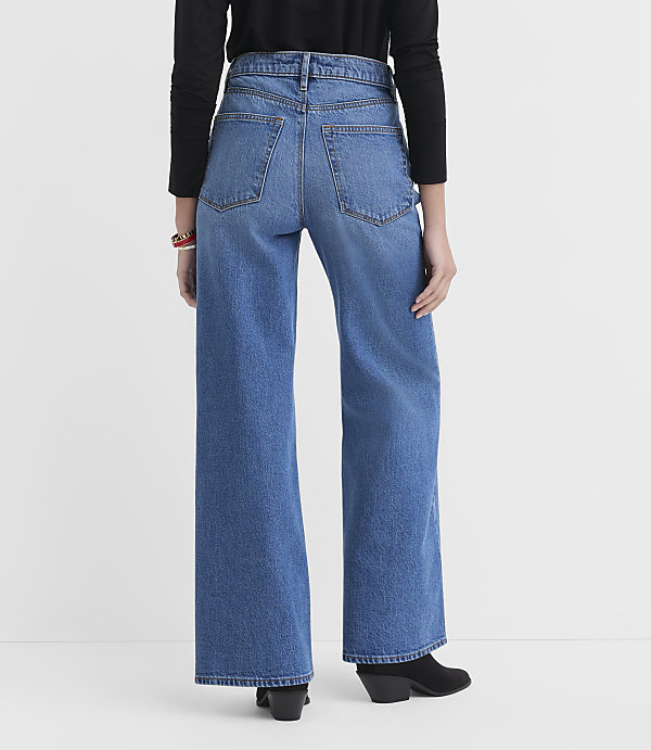 A person wearing wide-leg blue jeans and a black top stands against a plain white background.